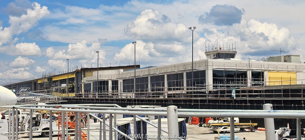 Asheville Regional Airport AVL Forward terminal construction in Fletcher near Asheville, North Carolina, viewed from the airport apron with scaffolding and exposed structural steel framing revealing the open terminal structure during construction. The airport terminal modernization and expansion was designed by Gresham Smith and is being built by Hensel Phelps for the Greater Asheville Regional Airport Authority – © 2026 David Oppenheimer – Performance Impressions photography archives https://performanceimpressions.com Asheville Regional Airport, Asheville Airport, Asheville Regional, Asheville airport terminal, Asheville terminal, Asheville airport construction, Asheville construction, Asheville airport expansion, AVL Forward, AVL Forward terminal, AVL Forward project, AVL terminal, AVL terminal construction, AVL construction, AVL airport, AVL, KAVL, airport terminal, terminal, terminal construction, airport construction, terminal expansion, airport expansion, terminal modernization, airport modernization, terminal renovation, new terminal, new airport terminal, airport building, terminal building, building construction, construction, construction site, construction project, construction progress, under construction, building under construction, unfinished building, unfinished structure, work in progress, scaffolding, scaffold, scaffolds, structural steel, steel structure, steel frame, steel framing, exposed steel, exposed structure, exposed framing, girders, girder, steel girders, beams, beam, steel beams, columns, steel columns, structural frame, building frame, open structure, open framing, building structure, structural construction, steel construction, construction detail, construction architecture, architecture, airport architecture, terminal architecture, aviation architecture, architectural construction, architectural engineering, engineering, structural engineering, civil engineering, infrastructure, airport infrastructure, aviation infrastructure, transportation infrastructure, infrastructure construction, commercial construction, large scale construction, capital project, airport development, airport improvement, terminal development, aviation construction, airside construction, airport operations, construction operations, airport apron, apron, aircraft apron, airport ramp, ramp, tarmac, airfield, airside, airport ground, ground boarding, passenger operations, aviation, airport, regional airport, commercial airport, transportation, air travel, Gresham Smith, Gresham Smith Asheville, Gresham Smith aviation, architect, architects, airport architect, terminal architect, aviation architect, architecture firm, Brad Sucher, structural engineer, civil engineer, engineering firm, interior design, airport planning, terminal planning, experiential design, wayfinding, Hensel Phelps, Hensel Phelps Construction, Hensel Phelps Asheville, construction manager, construction management, CM at Risk, contractor, general contractor, airport contractor, aviation contractor, Greater Asheville Regional Airport Authority, Asheville Regional Airport Authority, GARAA, airport authority, Asheville, Asheville North Carolina, North Carolina, NC, Western North Carolina, WNC, Fletcher, Fletcher North Carolina, Henderson County, Buncombe County, Blue Ridge, Blue Ridge Mountains, 61 Terminal Drive, Terminal Drive, Airport Road, 28732, United States, USA, airport photography, construction photography, architecture photography, architectural photography, infrastructure photography, aviation photography, transportation photography, industrial photography, documentary photography, editorial photography, commercial photography, Asheville photography, North Carolina photography, construction art, architecture art, airport art, Asheville art, photographic print, fine art print, archival print, gallery print, collector print, wall art, licensing, image licensing, commercial imagery, photography licensing, art, print, fine art, commercial photography, clouds, sunny, blue sky, summer, David Oppenheimer, Performance Impressions, Performance Impressions photography archives, horizontal, 1-15156784411