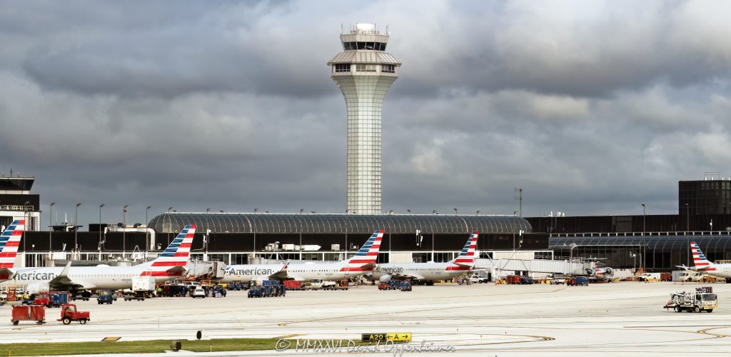 Chicago O’Hare International Airport Terminal 3 with American Airlines jets parked at the K gates beneath dark clouds, with the jet bridges, the airport apron, and an air traffic control tower in the background. The terminal section at the left edge houses the American Airlines flagship lounge, the Admirals Club – © 2026 David Oppenheimer – Performance Impressions photography archives https://performanceimpressions.com Chicago O’Hare International Airport, Chicago O’Hare Airport, O’Hare International Airport, O’Hare Airport, Chicago O’Hare, Chicago airport, ORD, O’Hare Terminal 3, Chicago O’Hare Terminal 3, Terminal 3, Terminal 3 Chicago, American Airlines jets, American Airlines O’Hare, American Airlines ORD, American Airlines Terminal 3, O’Hare K gates, Terminal 3 K gates, K gates, Concourse K, K Concourse, 10000 W O’Hare Avenue, 10000 West O’Hare Avenue, O’Hare, O’Hare Chicago, O’Hare community area, Northwest Side Chicago, Chicago Illinois, Chicago IL, 60666, Orchard Field, former Orchard Field, airport, terminal, airport terminal, gates, airport gates, boarding gates, gate area, aviation, commercial aviation, air travel, airline, airlines, jet, jets, aircraft, airplanes, airliners, passenger jets, passenger aircraft, commercial aircraft, parked jets, parked aircraft, aircraft parking, American Airlines, American, American Airlines Group, AAdvantage, Oneworld, American Airlines hub, airline hub, hub airport, American Airlines fleet, American Airlines livery, aircraft tails, jet bridge, jet bridges, apron, airport apron, ramp, airport ramp, tarmac, ground operations, airline operations, airport operations, airfield, terminal building, terminal exterior, airport exterior, building exterior, facade, air traffic control, air traffic control tower, airport control tower, control tower, ATC, ATC tower, O’Hare control tower, FAA, Federal Aviation Administration, Chicago Department of Aviation, CDA, American Airlines Flagship Lounge, American Flagship Lounge, Flagship Lounge, Flagship Lounge ORD, American Airlines Admirals Club, Admirals Club, Admirals Club ORD, Admirals Club lounge, American Airlines lounge, Terminal 3 lounge, H K connector, dark clouds, cloudy sky, overcast sky, cloud cover, weather, airport weather, aviation weather, dramatic sky, airport architecture, terminal architecture, transportation architecture, airport photography, aviation photography, transportation, travel, business travel, Chicago aviation, Illinois aviation, United States, licensing, image licensing, commercial imagery, photography licensing, art, print, fine art, commercial photography, David Oppenheimer, horizontal, 1-15213827141