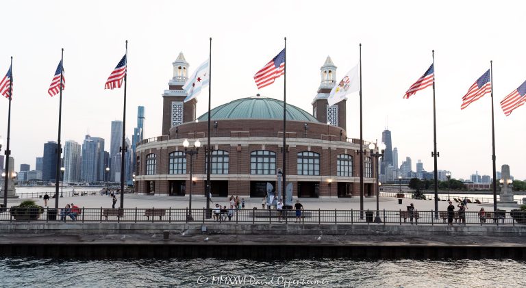 A postcard view of the Aon Grand Ballroom at Navy Pier from Lake Michigan, with the USS Chicago anchor, visitors along the seawall and nine flags flying against the Chicago skyline. Seven American flags join the Chicago and Illinois flags beneath a pale evening sky in Chicago, Illinois – © 2026 David Oppenheimer – Performance Impressions photography archives https://performanceimpressions.com Aon Grand Ballroom, 600 East Grand Avenue, Navy Pier, USS Chicago anchor, Chicago Warship Anchor, Chicago skyline, Chicago cityscape, Chicago postcard, Navy Pier postcard, Visit Chicago, Grand Ballroom, Navy Pier Auditorium, ballroom exterior, building exterior, facade, skyline, city skyline, cityscape, skyline view, Chicago skyline view, waterfront skyline, lakefront skyline, urban skyline, city view, lake view, postcard view, postcard image, postcard, Chicago waterfront, Chicago lakefront, Lake Michigan, flags, flag, American flag, American flags, U.S. flag, U.S. flags, USA flag, United States flag, Stars and Stripes, Chicago flag, Chicago city flag, city flag, red stars, Illinois flag, Illinois state flag, state flag, Illinois state seal, Illinois seal, flagpoles, flags flying, flags in wind, windblown flags, nine flags, seven American flags, waterfront promenade, seawall, visitors, tourists, people, evening, pale sky, Chicago tourism, Chicago travel, Chicago vacation, Chicago destination, Chicago attractions, Chicago landmarks, things to do in Chicago, tourist attraction, tourist destination, travel destination, sightseeing, tourism, travel, vacation, destination, iconic Chicago, Windy City, Illinois tourism, Midwest travel, Great Lakes tourism, Streeterville, Chicago, Illinois, Cook County, architecture, historic architecture, Chicago Landmark, Arts and Crafts architecture, twin towers, concert hall towers, tower lanterns, domed ballroom, Charles Sumner Frost, Frost and Granger, Edward C. Shankland, William Artingstall, Municipal Pier, Municipal Pier No. 2, Navy Pier Observation Deck, Offshore Rooftop, Offshore Rooftop and Bar, Bob Newhart Statue, Aon, Aon plc, Great Lakes, Midwest, USA, United States, 60611, travel photography, tourism photography, postcard photography, cityscape photography, destination imagery, landmark photography, Licensing, image licensing, commercial imagery, photography licensing, art, print, fine art, commercial photography, David Oppenheimer, horizontal, 1-15213827141