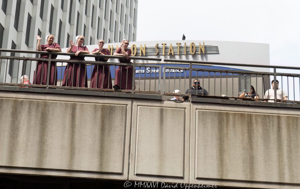 Women in matching burgundy dresses and white bonnet-style head coverings stand near Chicago Union Station by South Riverside Plaza, waving toward a boat tour on the Chicago River. The scene frames a candid downtown Chicago moment with bridge railings, riverfront architecture, and the Adams Street entrance area of the historic rail terminal – © 2026 David Oppenheimer – Performance Impressions photography archives https://performanceimpressions.com Chicago Union Station, Union Station, Union Station Chicago, women in burgundy dresses, burgundy dresses, white bonnets, bonnet headwear, white head coverings, coordinated outfits, matching dresses, costume-like clothing, theatrical style, Handmaid’s Tale style, dystopian style, public moment, urban event, unusual sighting, boat tour, Chicago boat tour, river tour, tour boat, Chicago River, river, riverfront, waterfront, water, bridge railing, metal railing, weathered railing, South Riverside Plaza, Riverside Plaza, Adams Street entrance, Union Station Adams Street, Welcome to Chicago, West Loop, Near West Side, downtown Chicago, Chicago Illinois, Chicago IL, 60606, rail station, train station, railroad station, transportation, transit, Amtrak, Metra, architecture, architectural facade, facade, building exterior, exterior, Beaux Arts, Beaux-Arts architecture, Classical Revival, historic building, landmark building, Chicago Landmark, Graham Anderson Probst White, Graham Anderson Probst & White, Daniel Burnham, Chicago architecture, historic architecture, urban photography, street photography, documentary photography, people, women, group, waving, posing, summer daylight, city tour, tourism, travel, United States, licensing, image licensing, commercial imagery, photography licensing, art, print, fine art, commercial photography, David Oppenheimer, horizontal, 1-15156784411