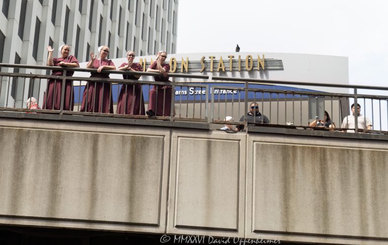 Women in matching burgundy dresses and white bonnet-style head coverings stand near Chicago Union Station by South Riverside Plaza, waving toward a boat tour on the Chicago River. The scene frames a candid downtown Chicago moment with bridge railings, riverfront architecture, and the Adams Street entrance area of the historic rail terminal – © 2026 David Oppenheimer – Performance Impressions photography archives https://performanceimpressions.com Chicago Union Station, Union Station, Union Station Chicago, women in burgundy dresses, burgundy dresses, white bonnets, bonnet headwear, white head coverings, coordinated outfits, matching dresses, costume-like clothing, theatrical style, Handmaid’s Tale style, dystopian style, public moment, urban event, unusual sighting, boat tour, Chicago boat tour, river tour, tour boat, Chicago River, river, riverfront, waterfront, water, bridge railing, metal railing, weathered railing, South Riverside Plaza, Riverside Plaza, Adams Street entrance, Union Station Adams Street, Welcome to Chicago, West Loop, Near West Side, downtown Chicago, Chicago Illinois, Chicago IL, 60606, rail station, train station, railroad station, transportation, transit, Amtrak, Metra, architecture, architectural facade, facade, building exterior, exterior, Beaux Arts, Beaux-Arts architecture, Classical Revival, historic building, landmark building, Chicago Landmark, Graham Anderson Probst White, Graham Anderson Probst & White, Daniel Burnham, Chicago architecture, historic architecture, urban photography, street photography, documentary photography, people, women, group, waving, posing, summer daylight, city tour, tourism, travel, United States, licensing, image licensing, commercial imagery, photography licensing, art, print, fine art, commercial photography, David Oppenheimer, horizontal, 1-15156784411